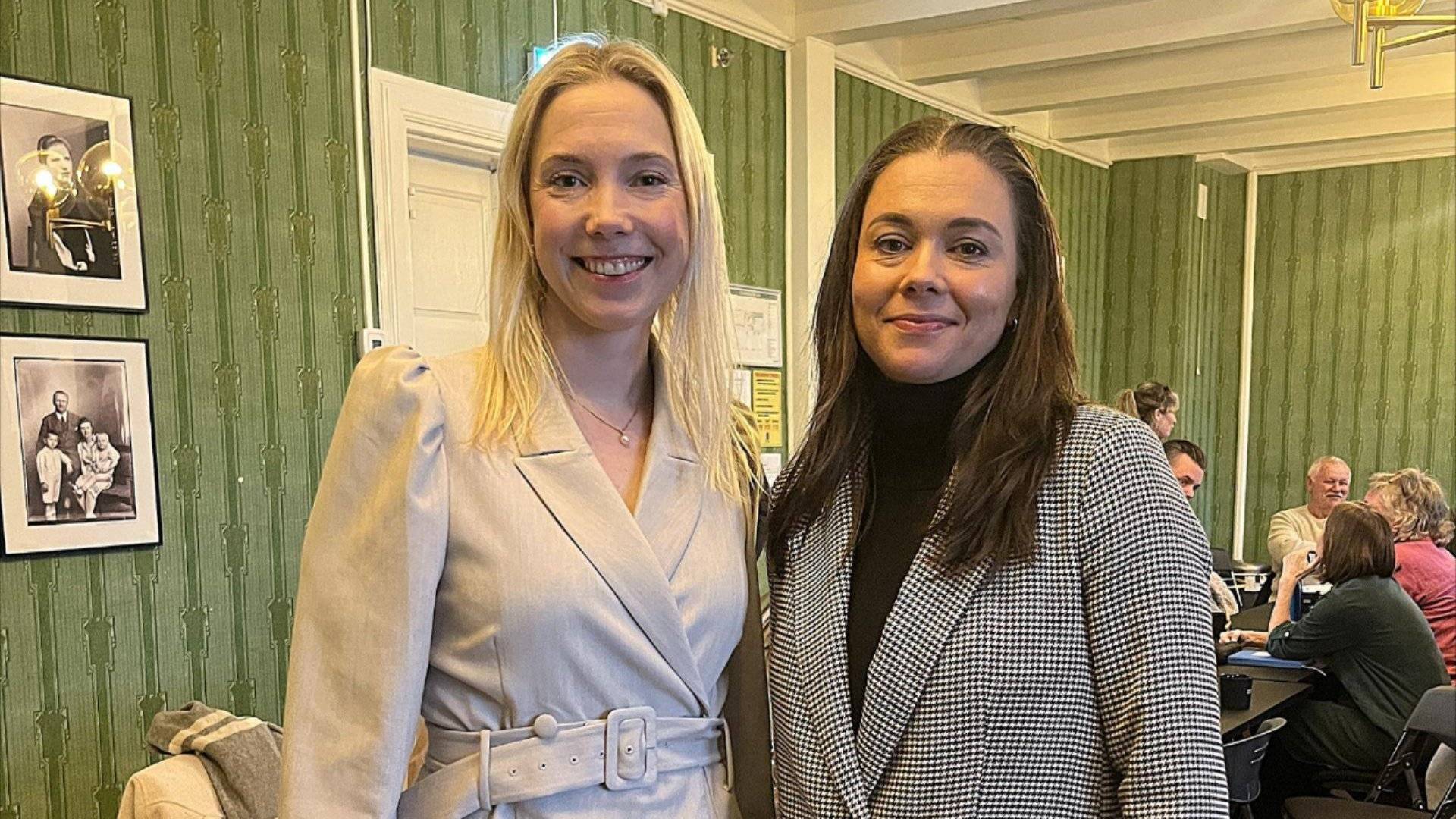 Oppstilt bilde av Hege Julianne Nevestad og Stine Karin Håtuft inne i et møterom. Mennesker i bakgrunnen.